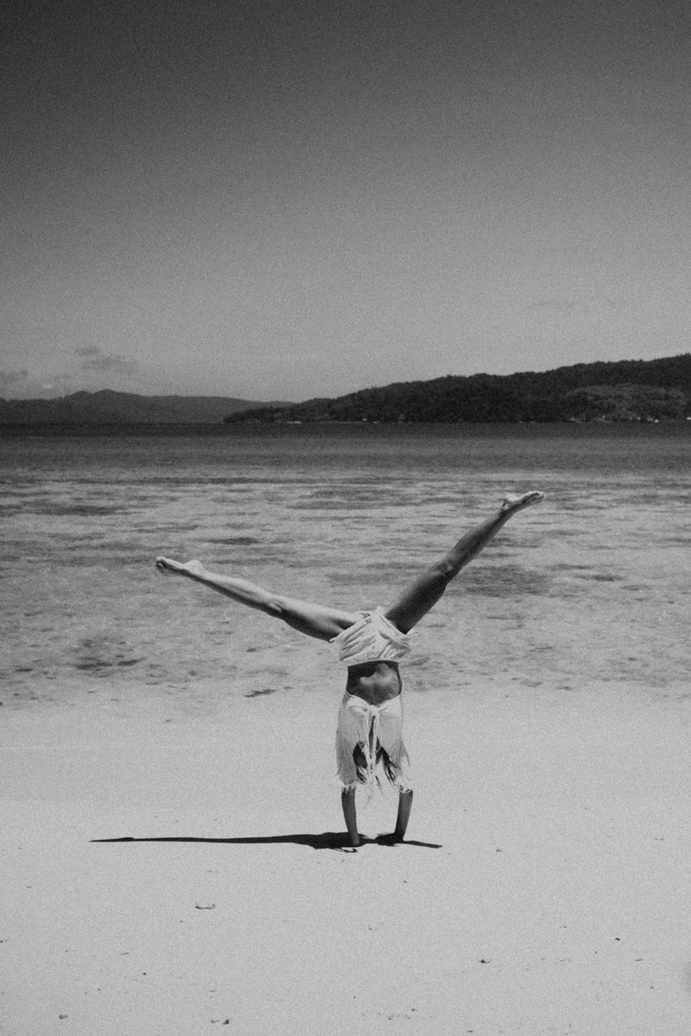 A woman balances in a handstand on a quiet beach, framed by calm water and distant hills beneath an open sky, creating a striking sense of space, balance, and simplicity.