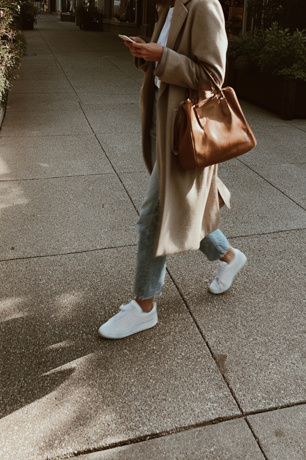 Woman in a neutral coat walking through a city while checking her phone, representing an intentional approach to building and growing a small business.