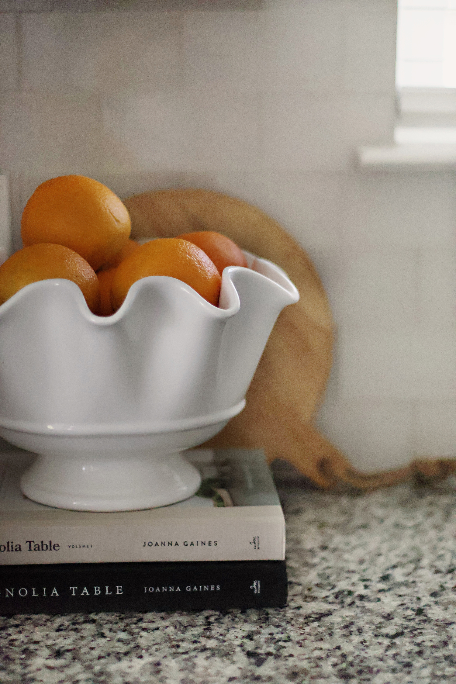 White ceramic bowl filled with fresh oranges styled on cookbooks in a bright kitchen.