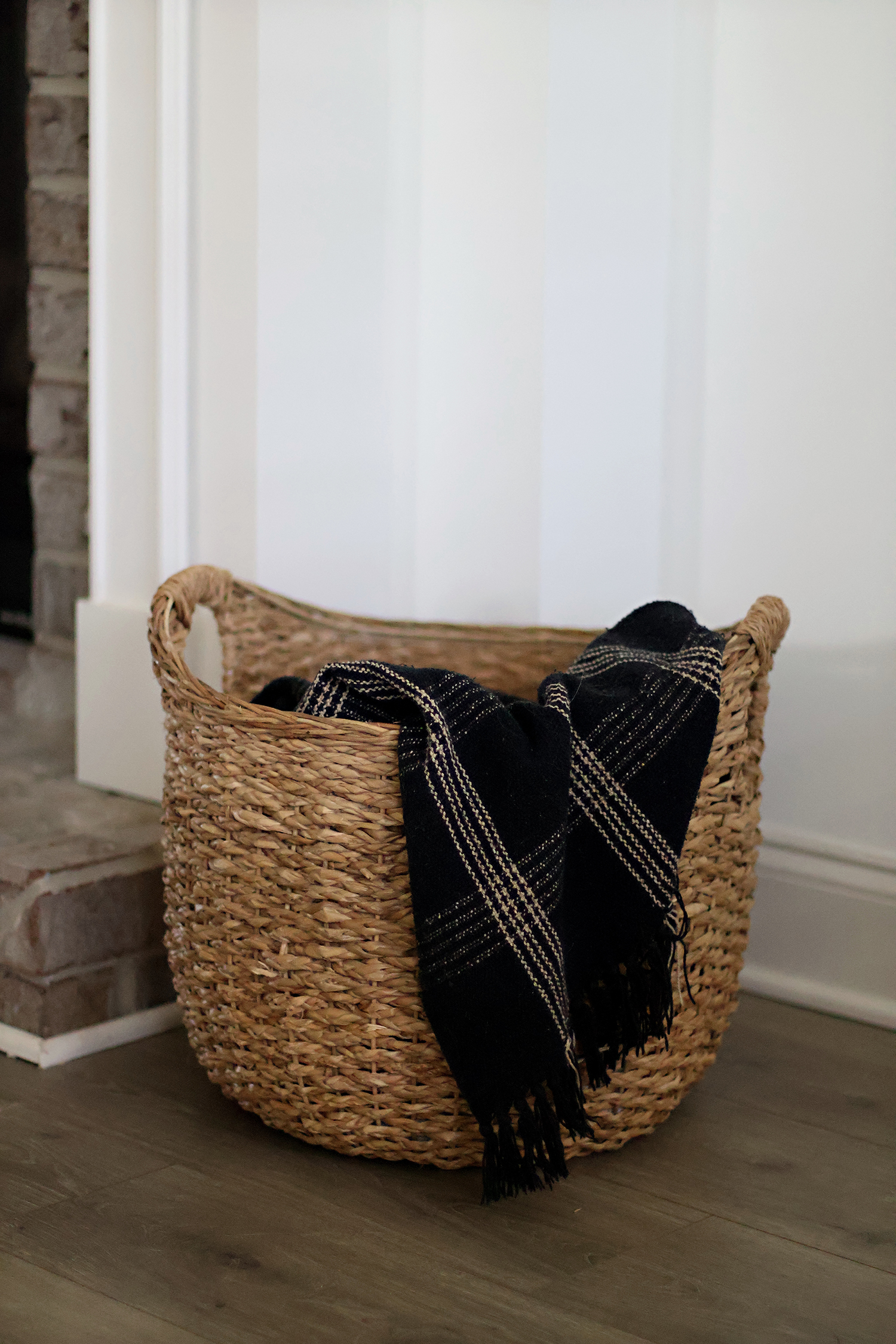 Natural woven storage basket holding a folded throw blanket beside a fireplace.